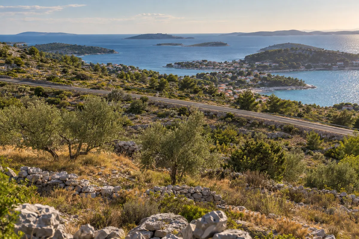 The coastal road crosses a dry Dalmatian hillside above an island-fringed bay and a small settlement.