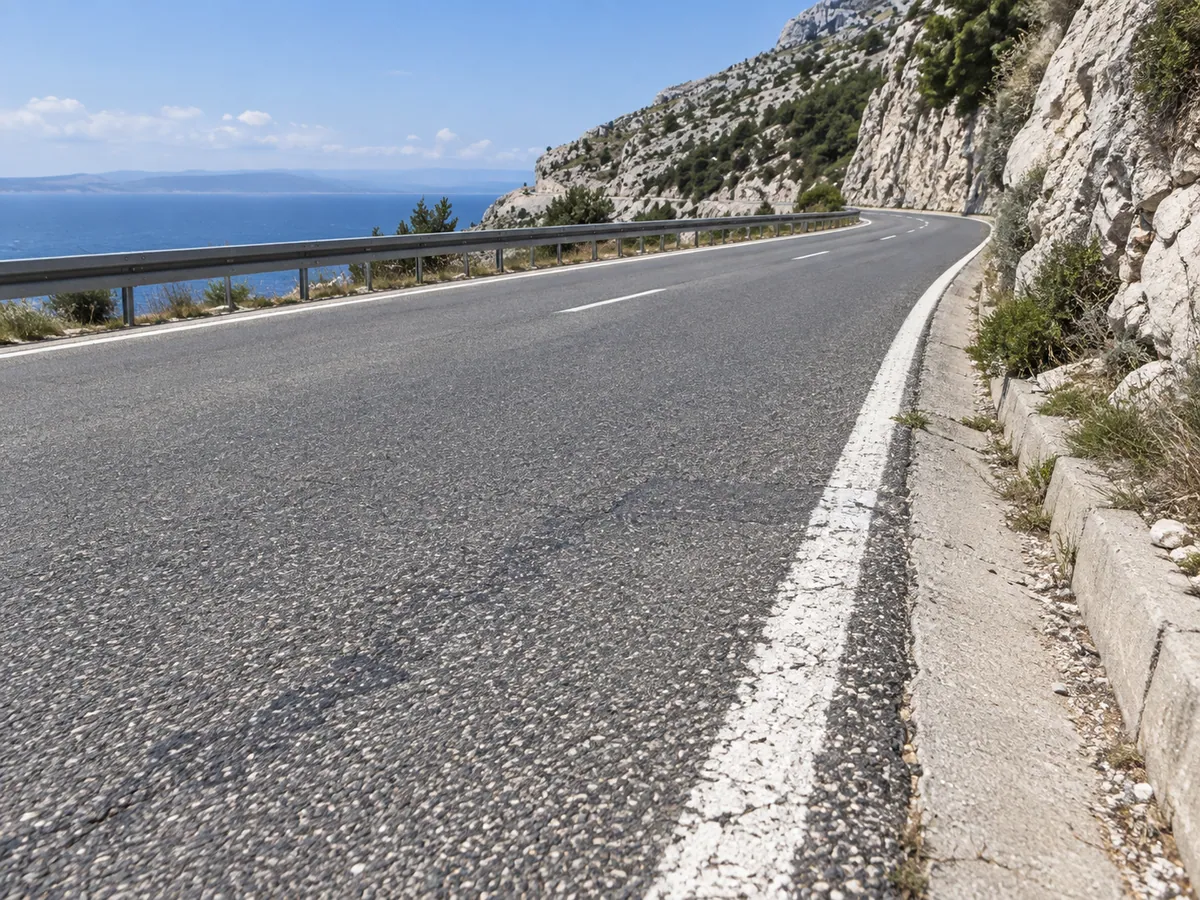 Coarse grey asphalt, a weathered white edge line and a drainage gutter run beside pale limestone.