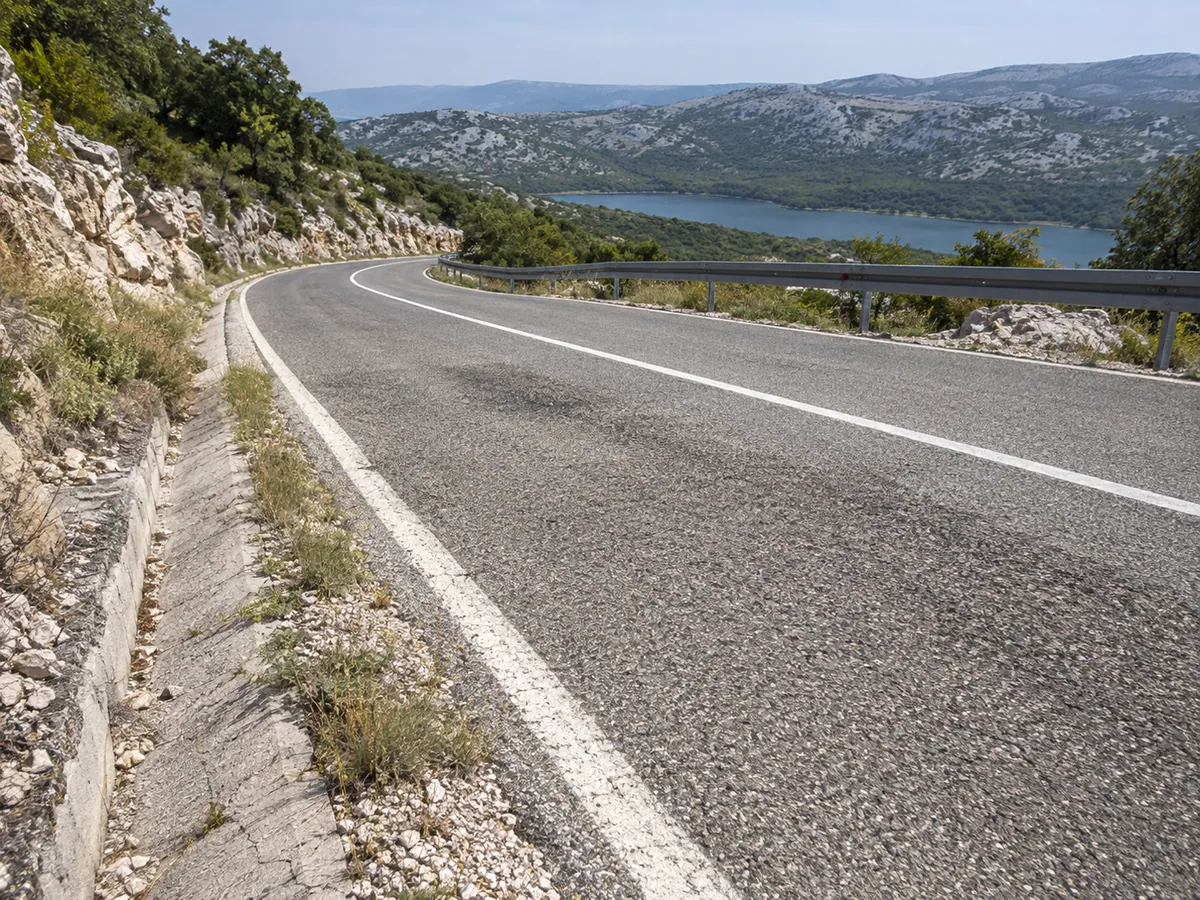 A place-inspired D100 bend shows coarse asphalt, road markings, drainage, and a narrow edge in central Cres.