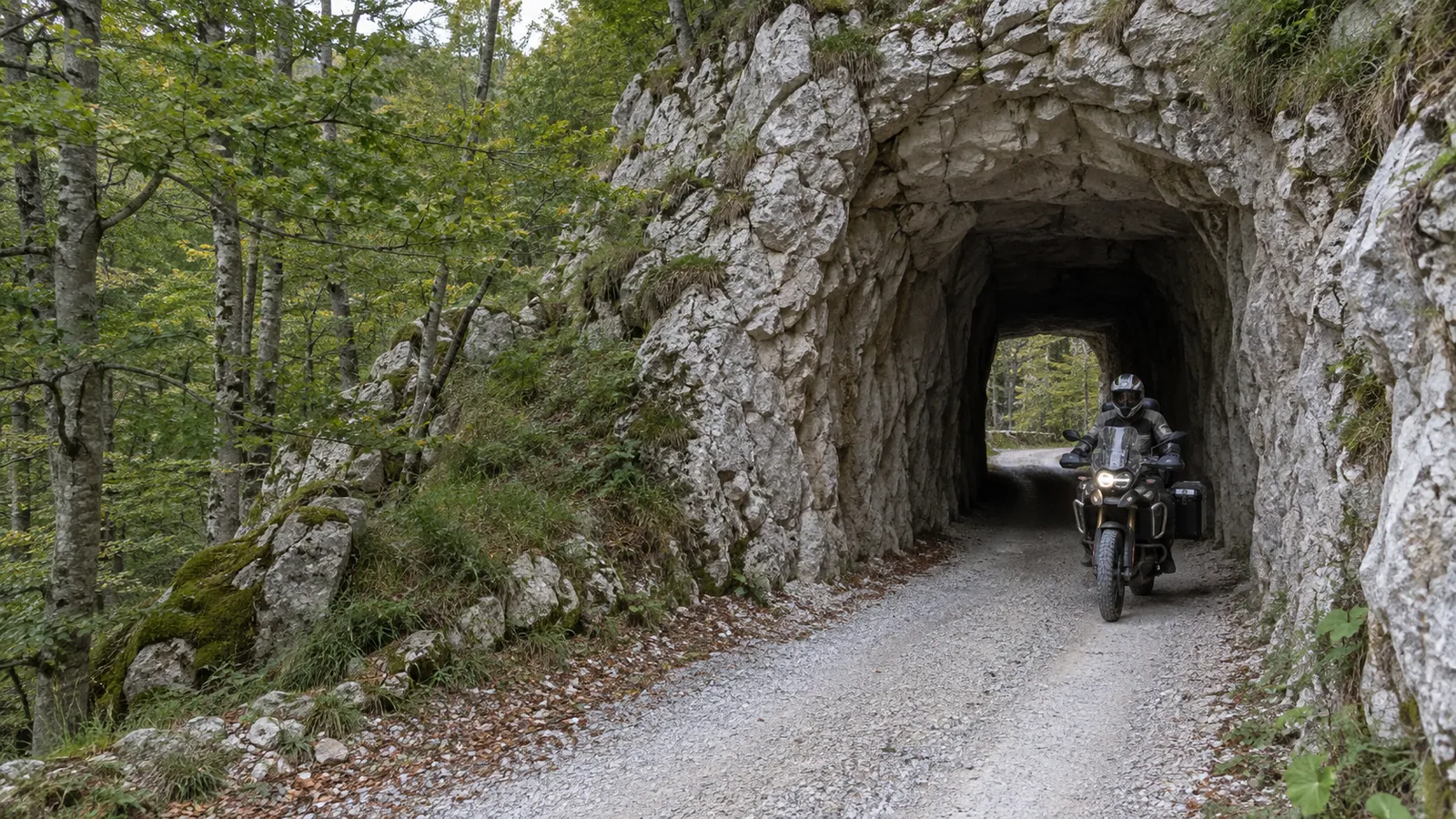 An adventure motorcycle exits a short rock tunnel on the pale gravel Dabarska Road beneath Velebit forest.