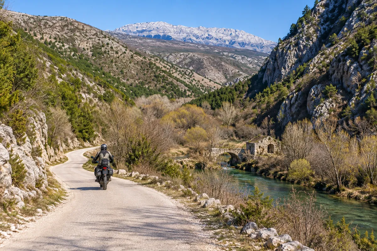 An adventure rider follows a gravel road beside the green Krčić corridor and low limestone slopes.