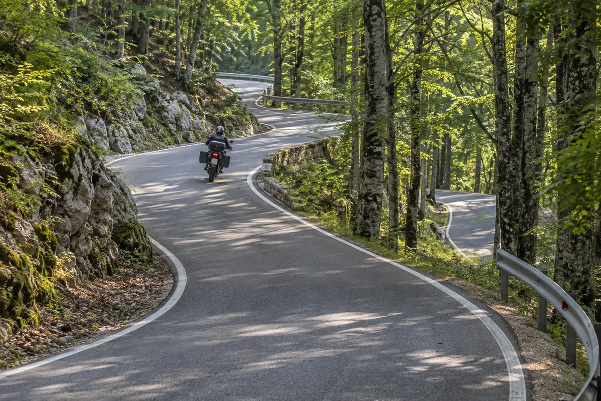 A touring rider climbs through closely spaced shaded bends on the forested public road toward Poklon Pass.